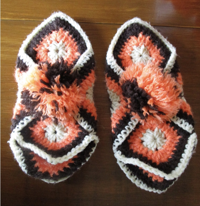 Two crocheted slippers in white, brown, and orange yarn with a large pom pom in the center of each.